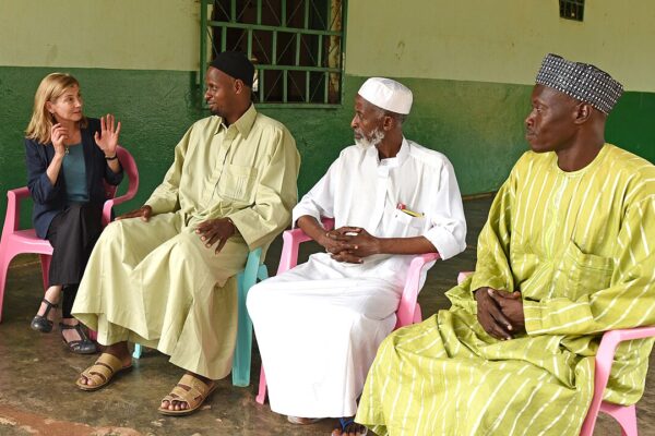 Three Muslim leaders in robes and a woman in a blazer sit on plastic chairs on a veranda in Bangui, talking