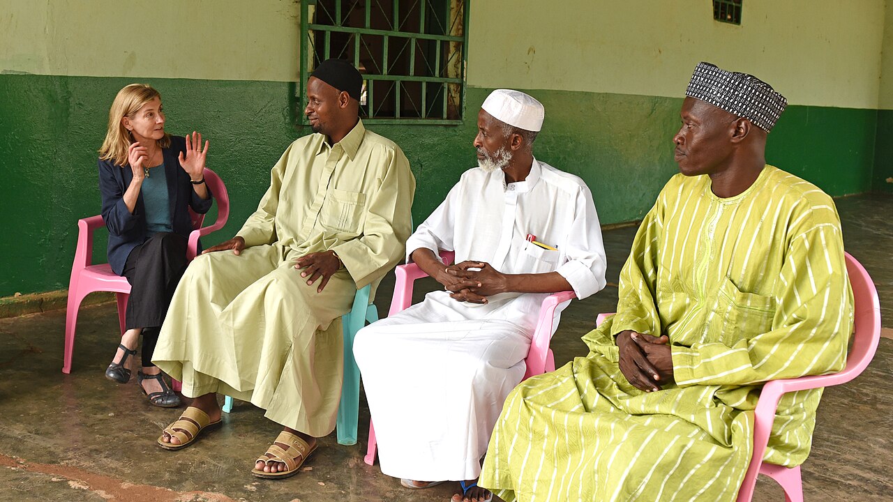 Three men in long robes and a woman in a blazer sit on plastic chairs in a covered yard in Bangui, talking