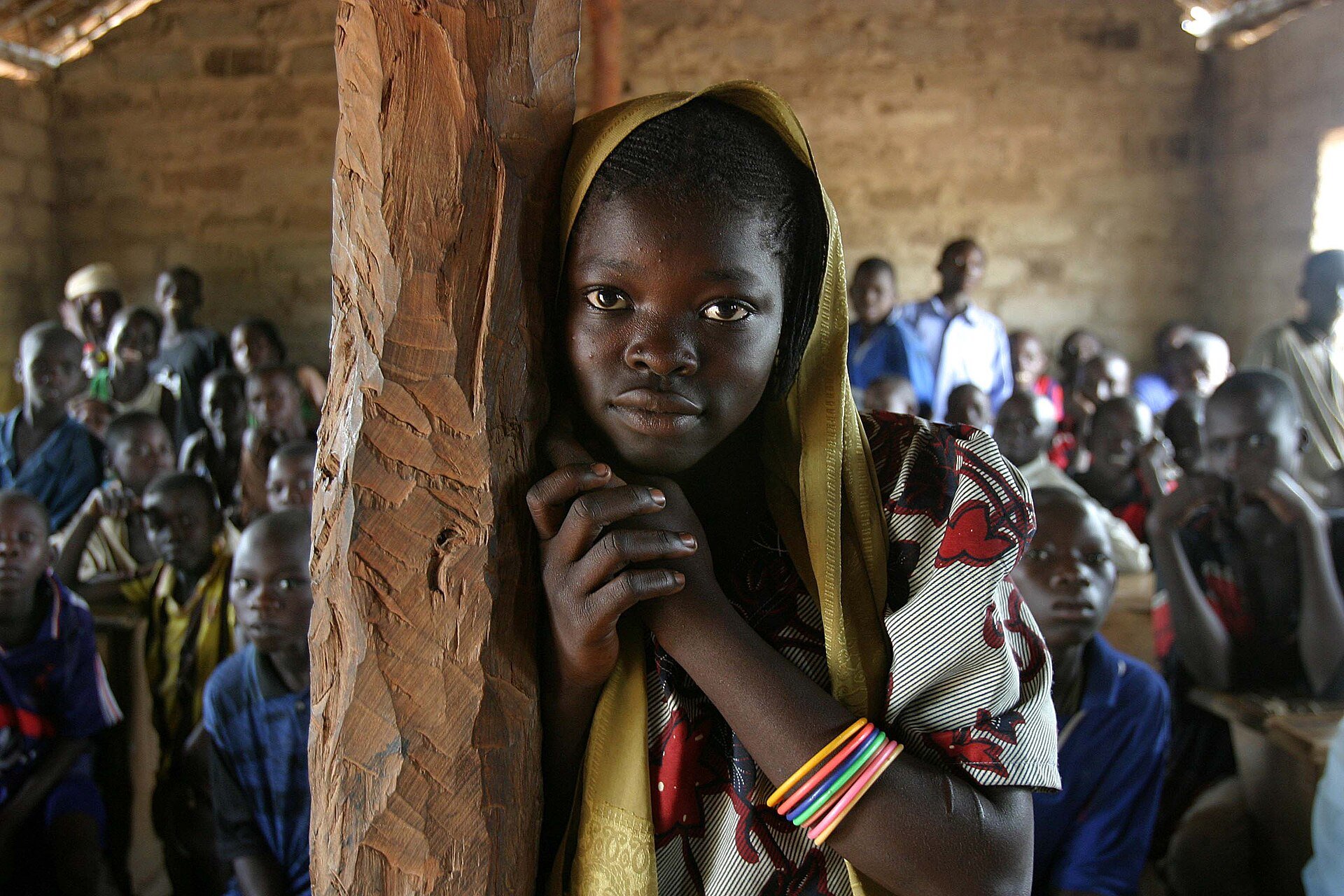 A pupil in a yellow headscarf leans on a carved wooden post in a crowded classroom in the Central African Republic, with pupils seated behind her