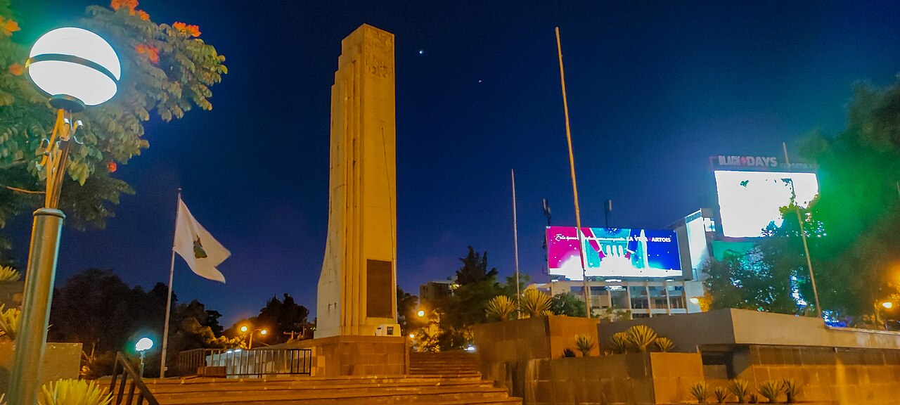 The illuminated stone Obelisk in Guatemala City at night with a white flag beside it