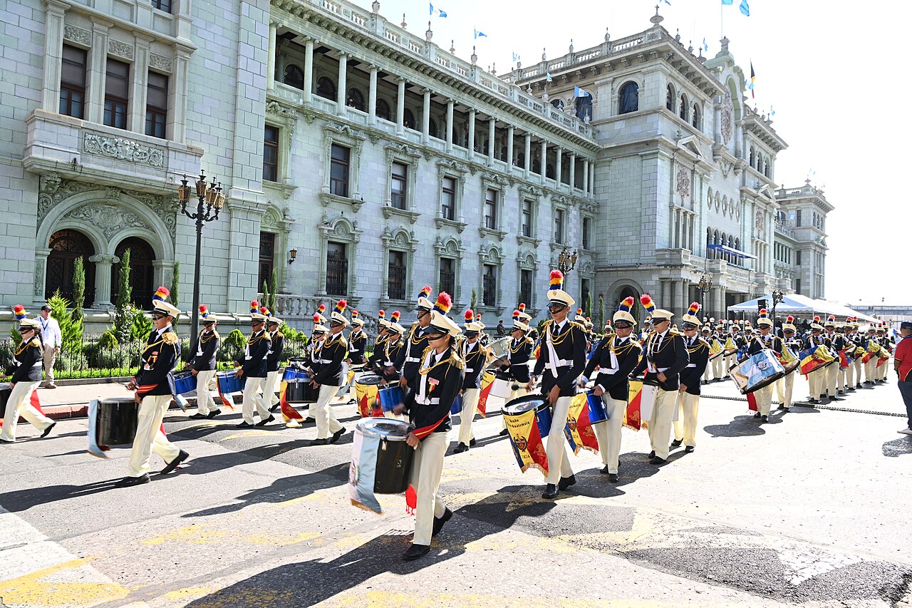 School drummers in dark uniforms and plumed helmets marching past the National Palace of Culture in Guatemala City