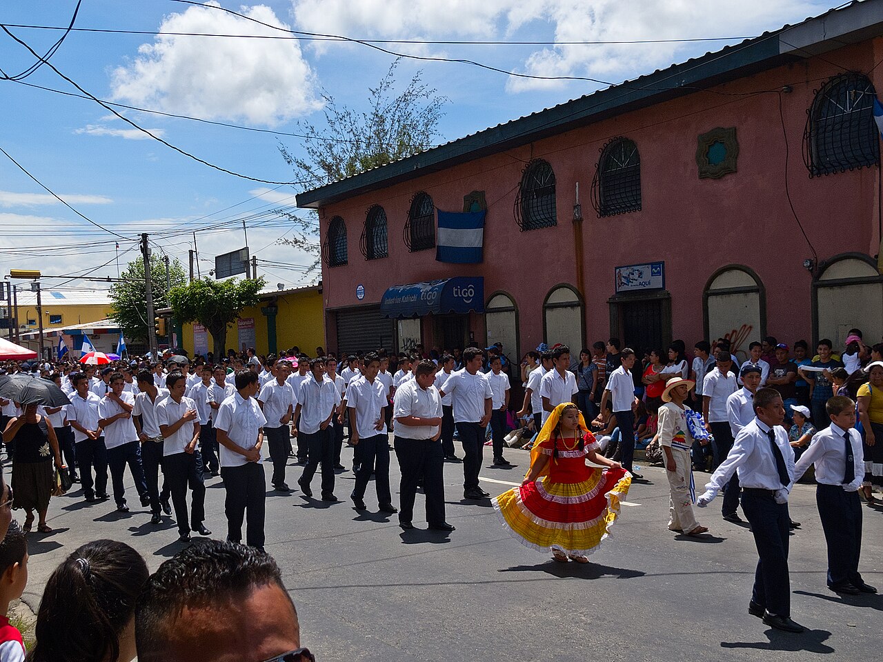 Students in white shirts march and a girl in a red and yellow folk dress dances in an independence parade in Santa Tecla, El Salvador