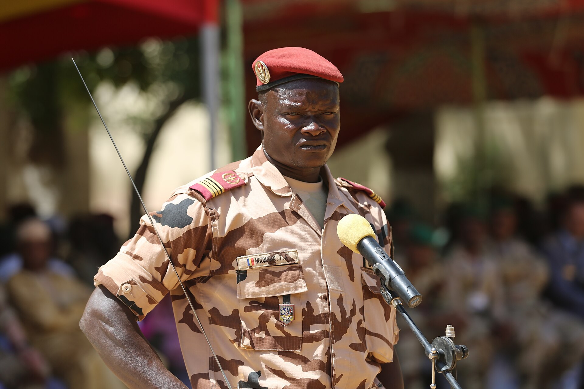 A Chadian Army soldier in desert camouflage and a red beret speaking into a microphone at a military ceremony in N'Djamena, Chad