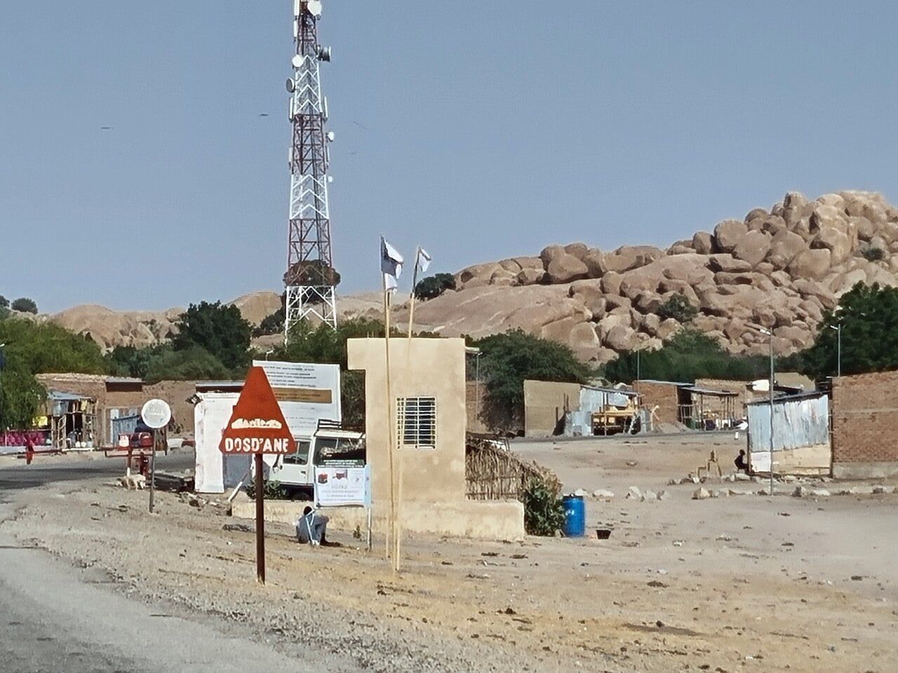 The entrance of Ngoura in Chad's Hadjer-Lamis province, with a mobile phone mast, a road sign and rocky hills behind roadside shops