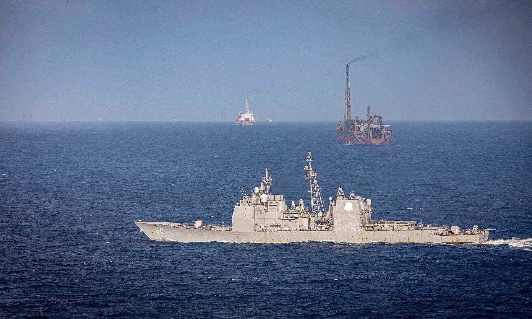 A US Navy cruiser passes a floating production vessel with a flare in the Stabroek Block off Guyana