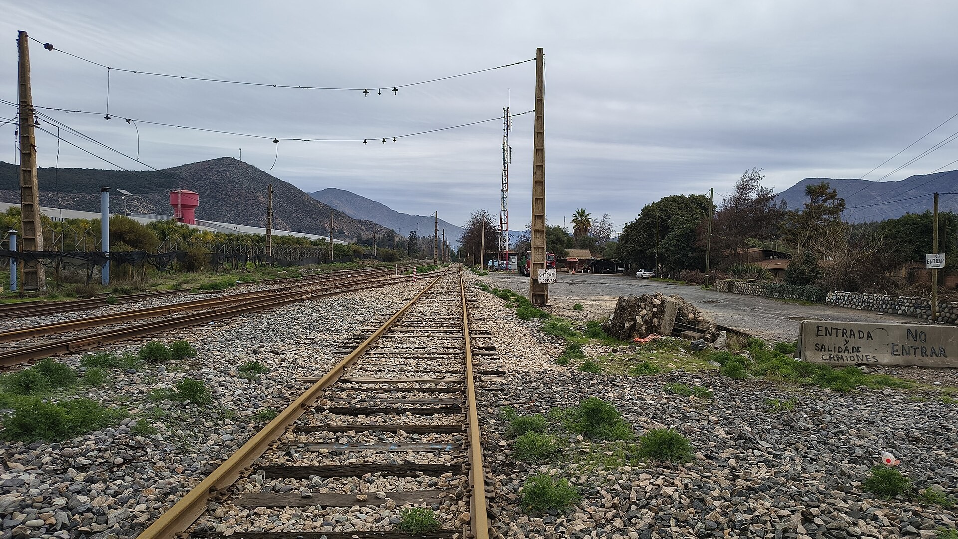 Railway tracks, power poles and a red water tower beside the Chagres smelter yard in Chile, with hills under a cloudy sky