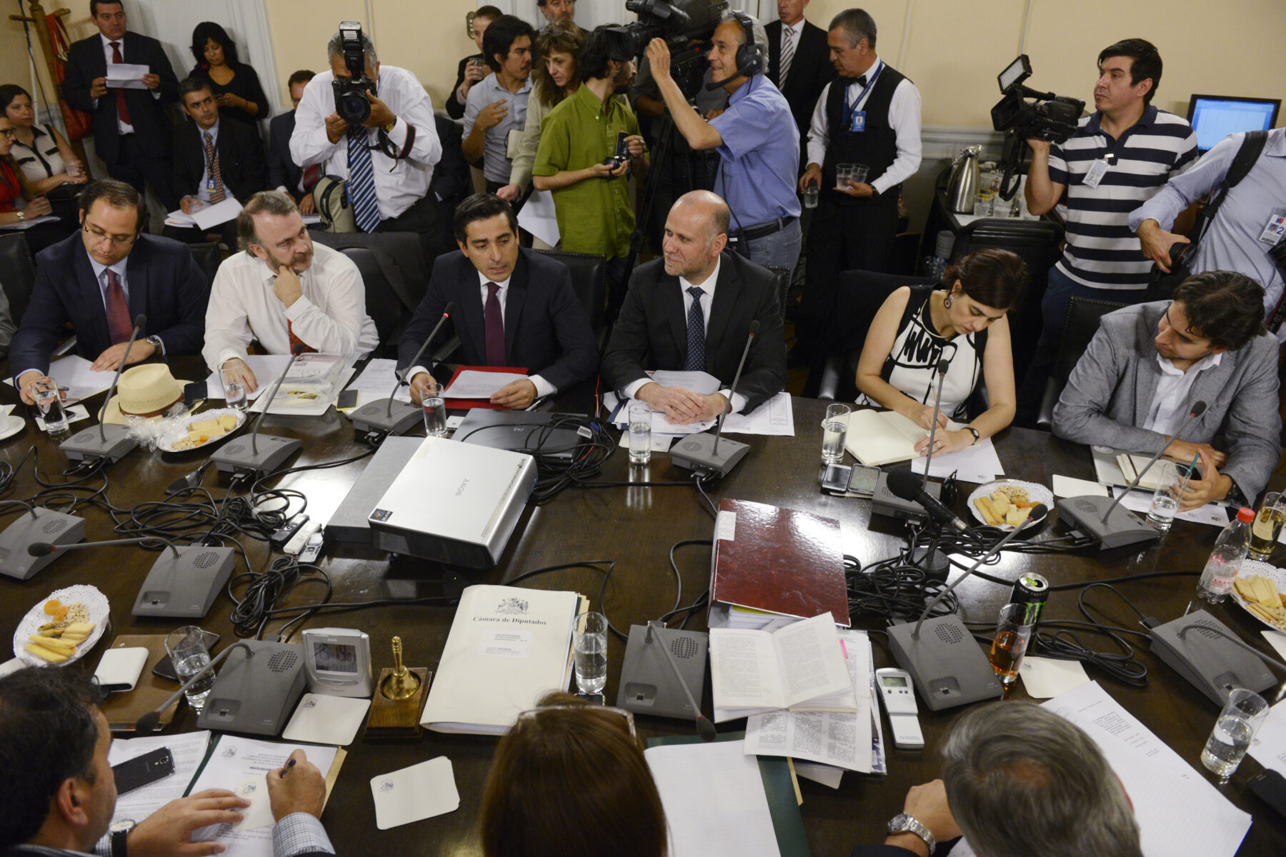 A Chilean Chamber of Deputies investigative committee in session, with deputies and a minister seated around a table and press cameras behind