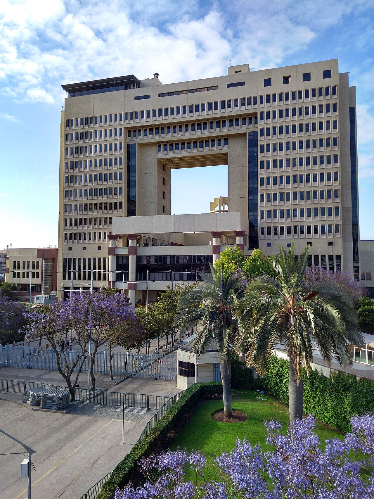 The National Congress building in Valparaíso, Chile, a tall block with a square opening, with palm trees and flowering jacaranda trees in front