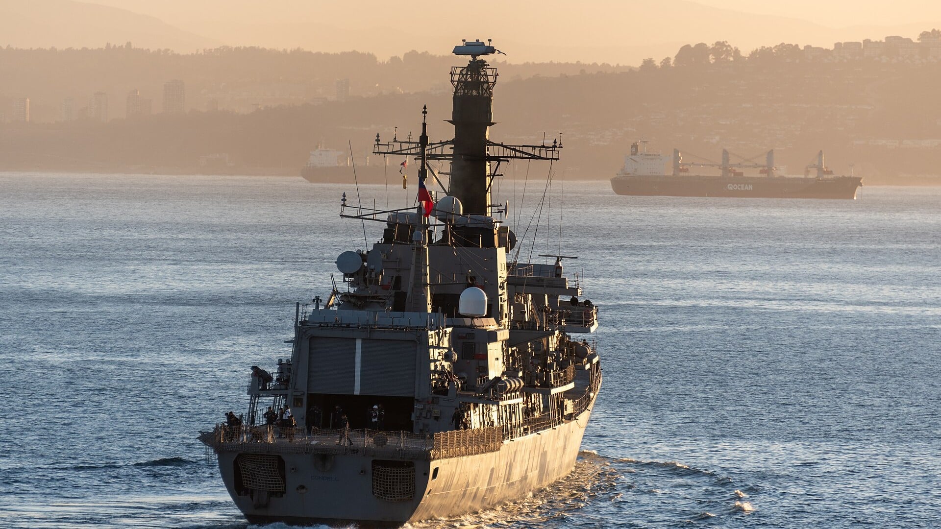 A Chilean Navy frigate seen from astern in warm low light as it leaves Valparaíso harbour, with a cargo ship at anchor behind it