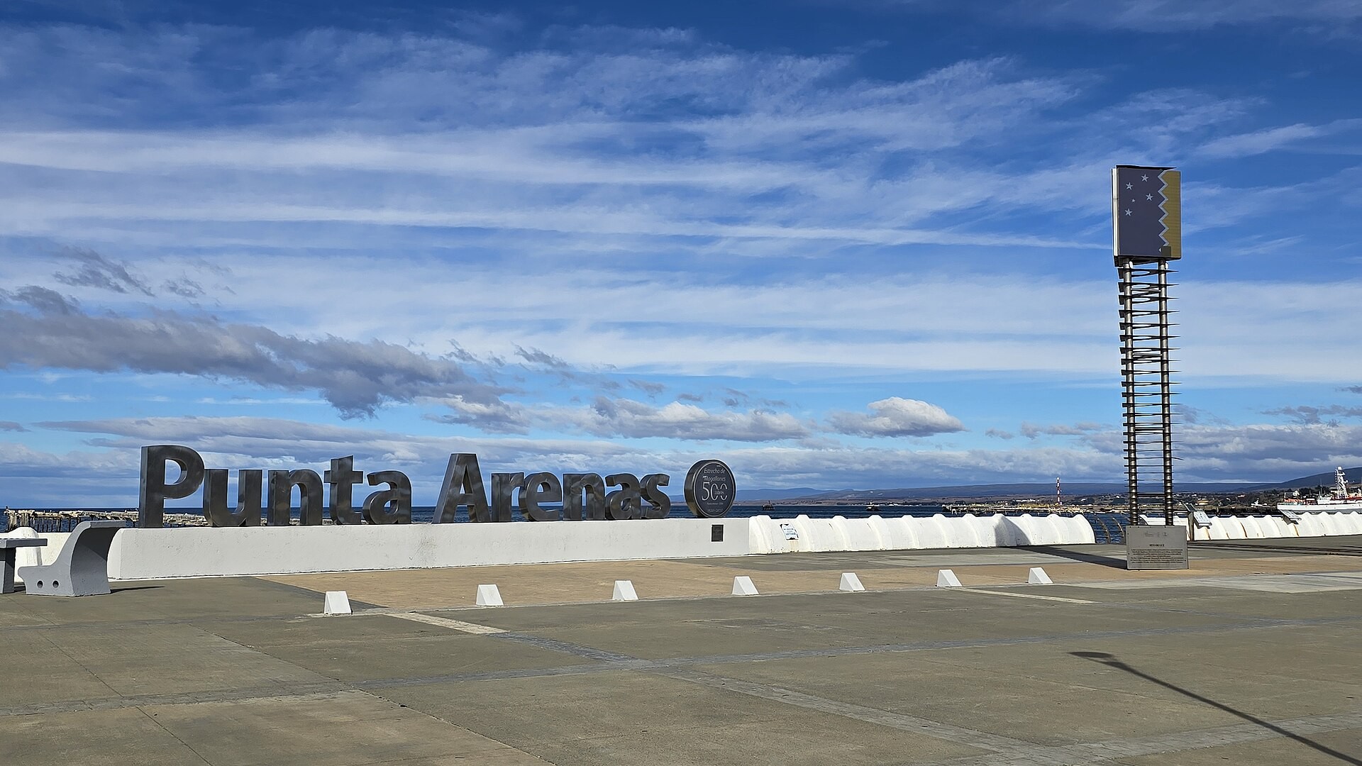 The Punta Arenas sign and a tall marker on the paved waterfront beside the Strait of Magellan under a blue sky with thin clouds