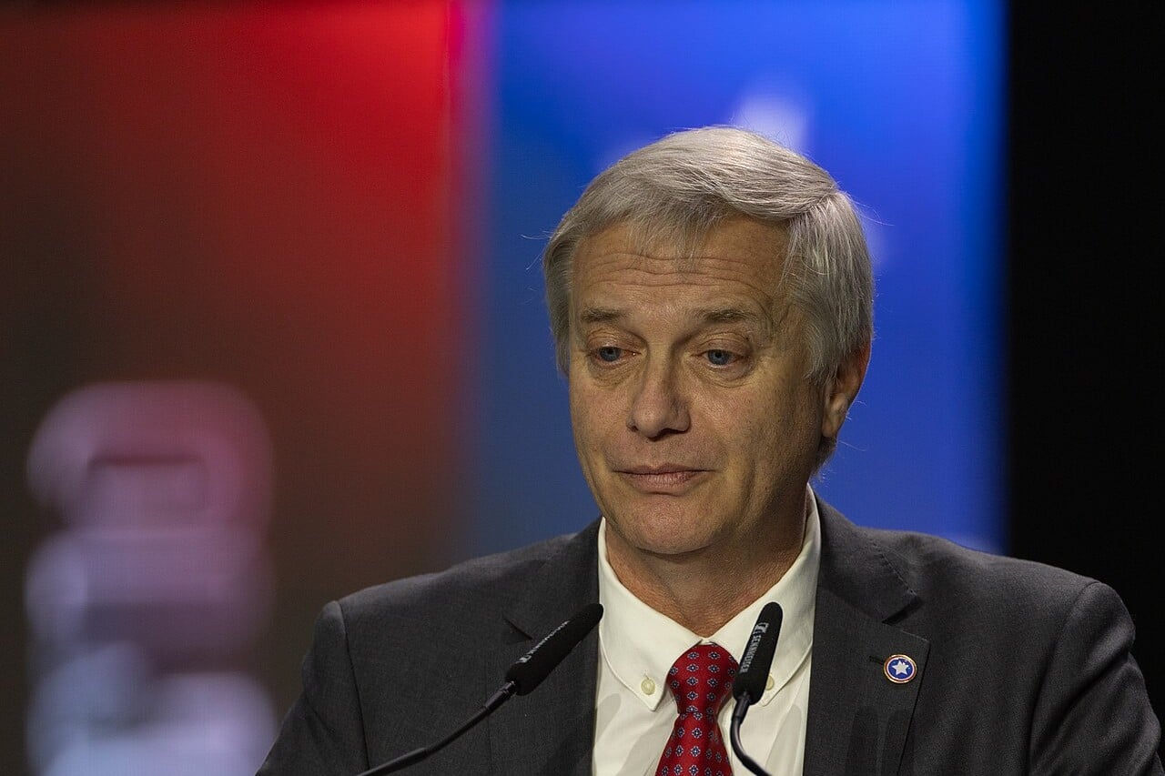 José Antonio Kast, grey-haired in a dark suit and red tie, speaking at a lectern with two microphones