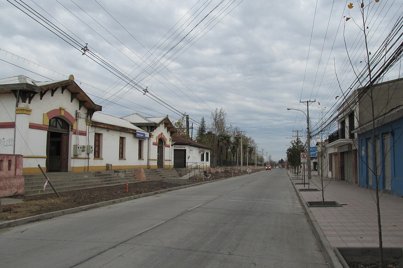 A wide street under overcast skies in Requínoa, with a low white building with a tiled roof on the left and shops on the right