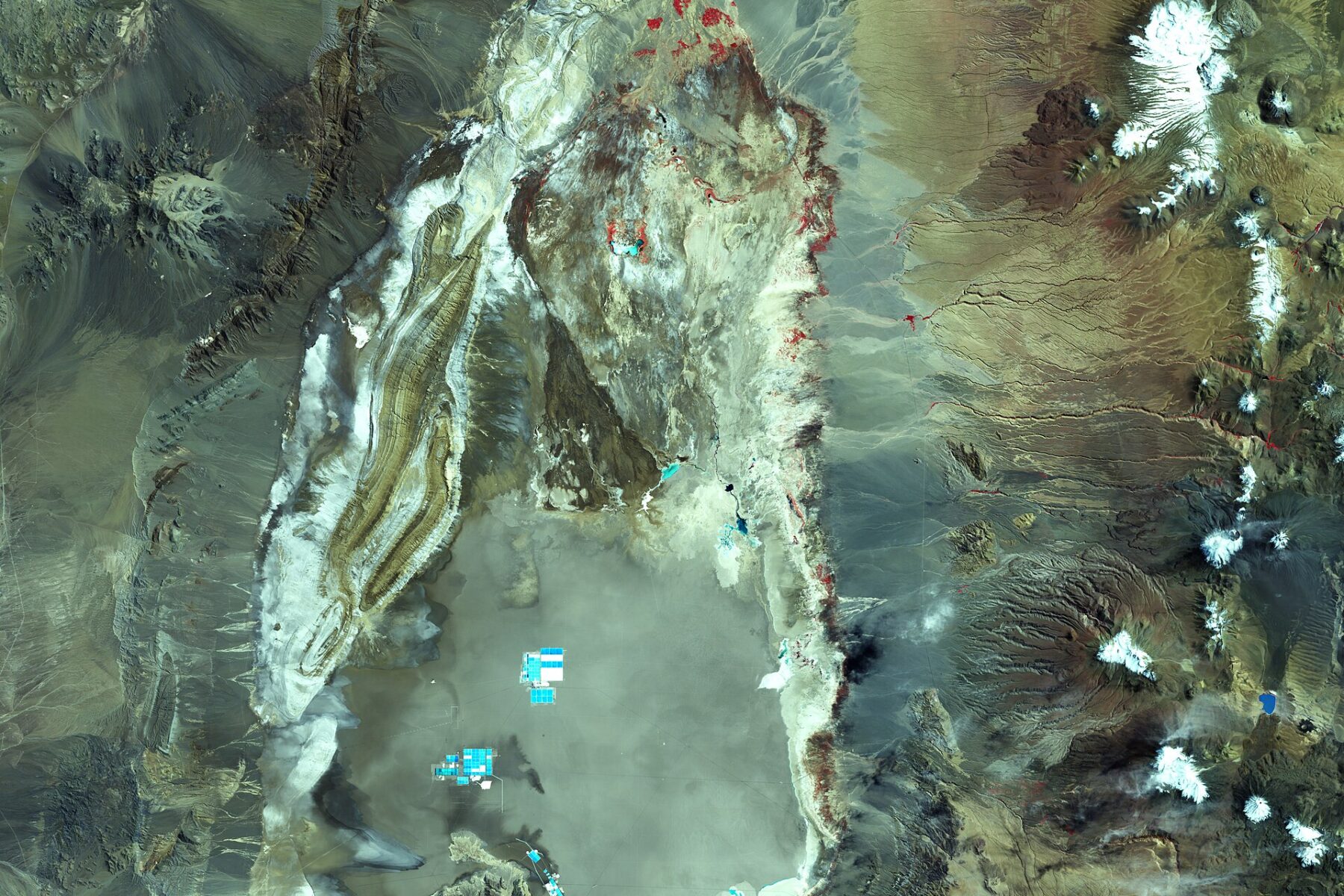 Satellite view of the Salar de Atacama salt flat in northern Chile, with blue lithium evaporation ponds visible and snow-capped volcanoes to the east