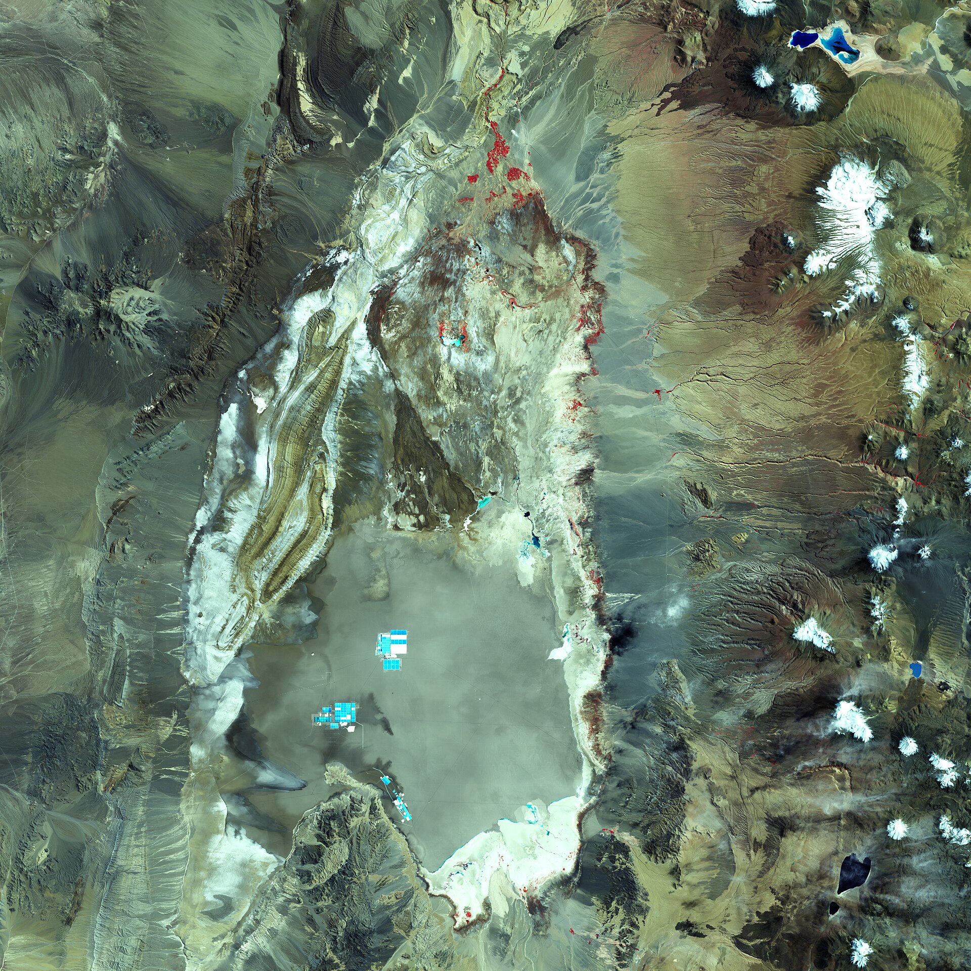 Satellite view of the Salar de Atacama salt flat in northern Chile, with blue lithium evaporation ponds visible and snow-capped volcanoes to the east