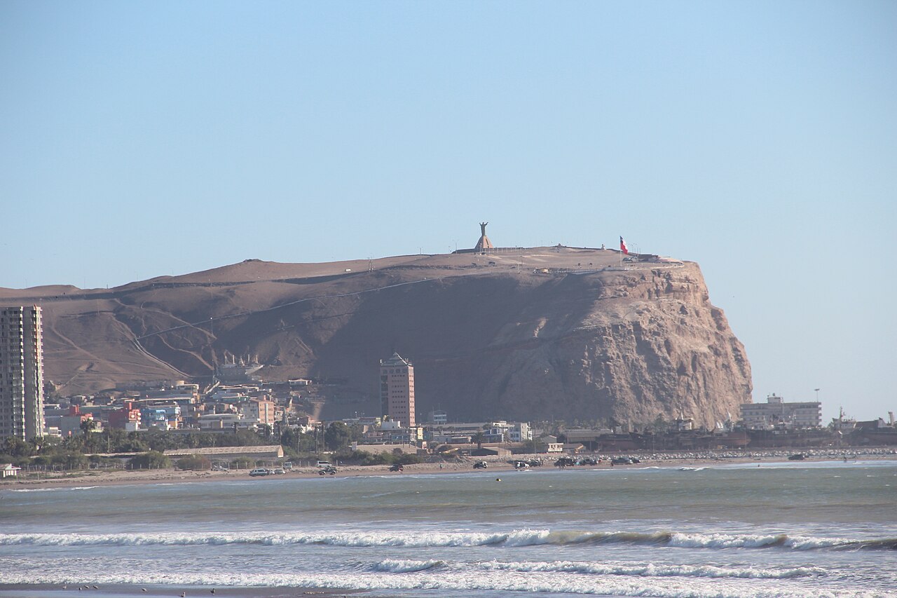 The Morro de Arica, a steep brown headland topped by a Chilean flag and a monument, rising behind the town and a beach with breaking waves