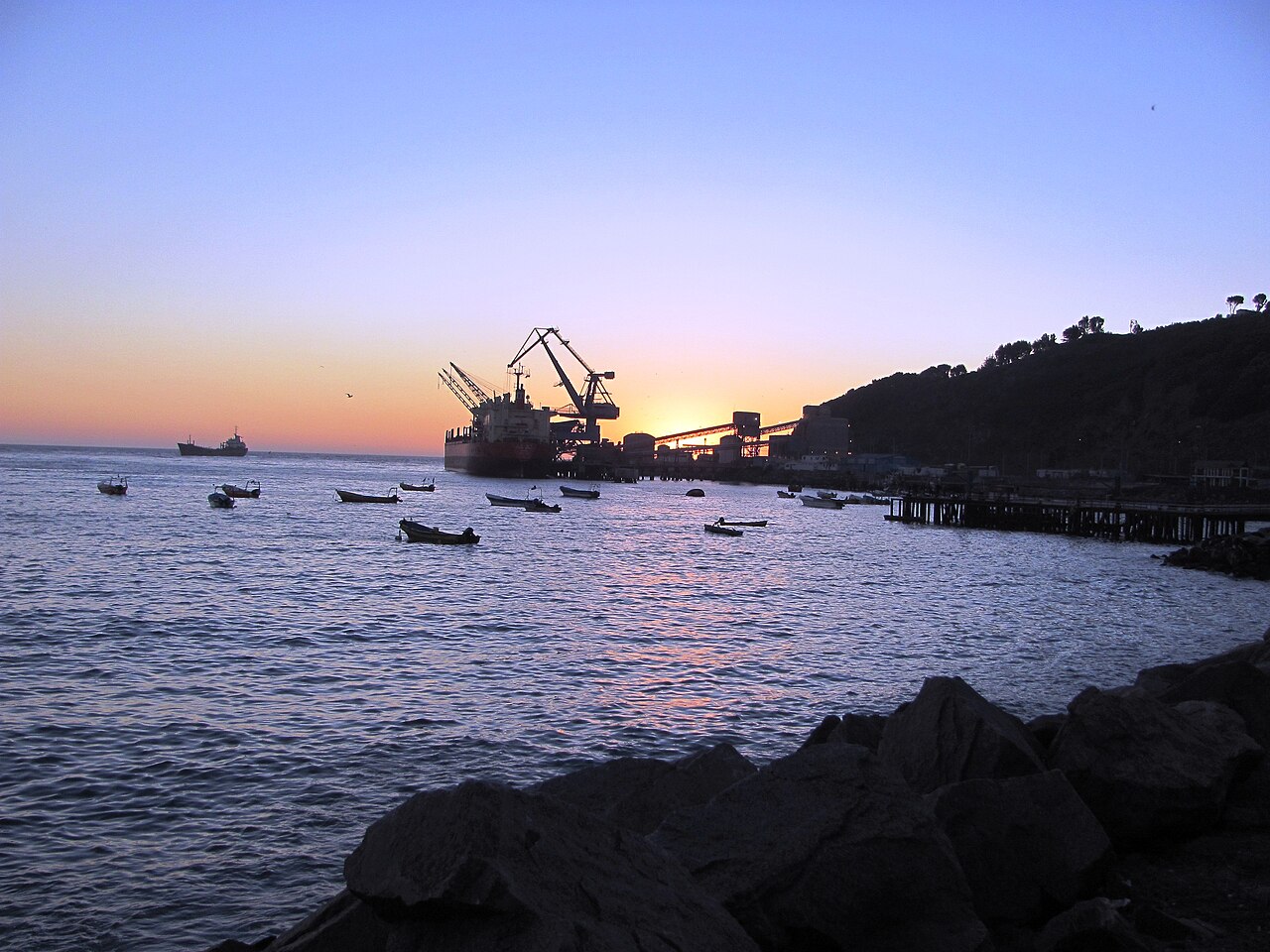 A ship moored at a pier under cranes in San Antonio harbour at dusk, with small boats on the water