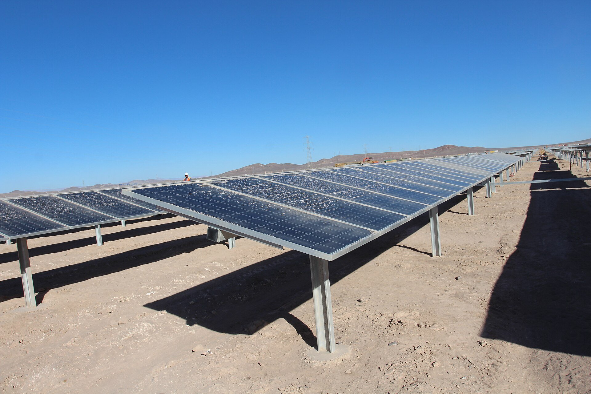 Rows of dusty solar panels on bare desert ground under a clear blue sky in northern Chile