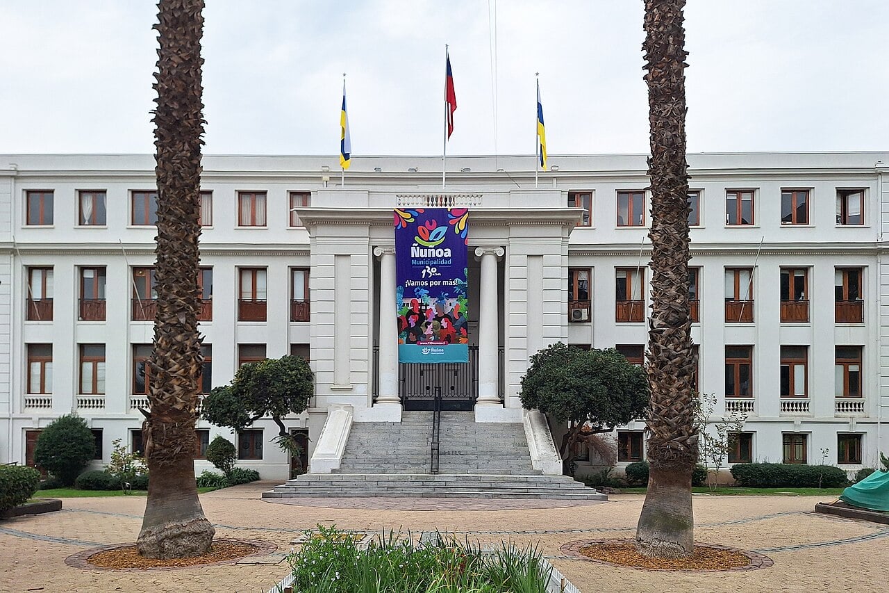 White neoclassical town hall of Ñuñoa with a colourful municipal banner over the entrance, flags on the roof and two palm trees