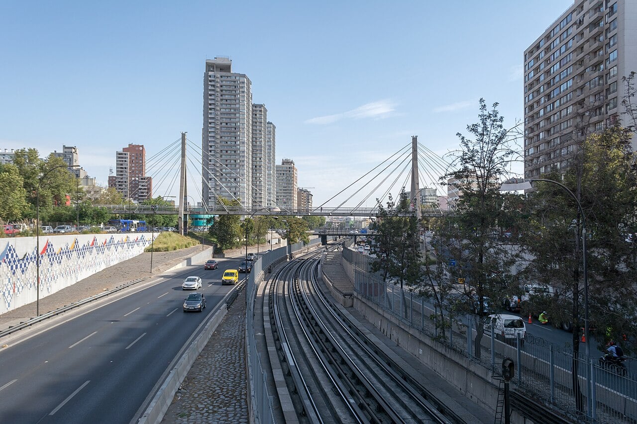 Autopista Central running beside Santiago Metro tracks under a cable-stayed footbridge, with cars on the highway and apartment towers behind