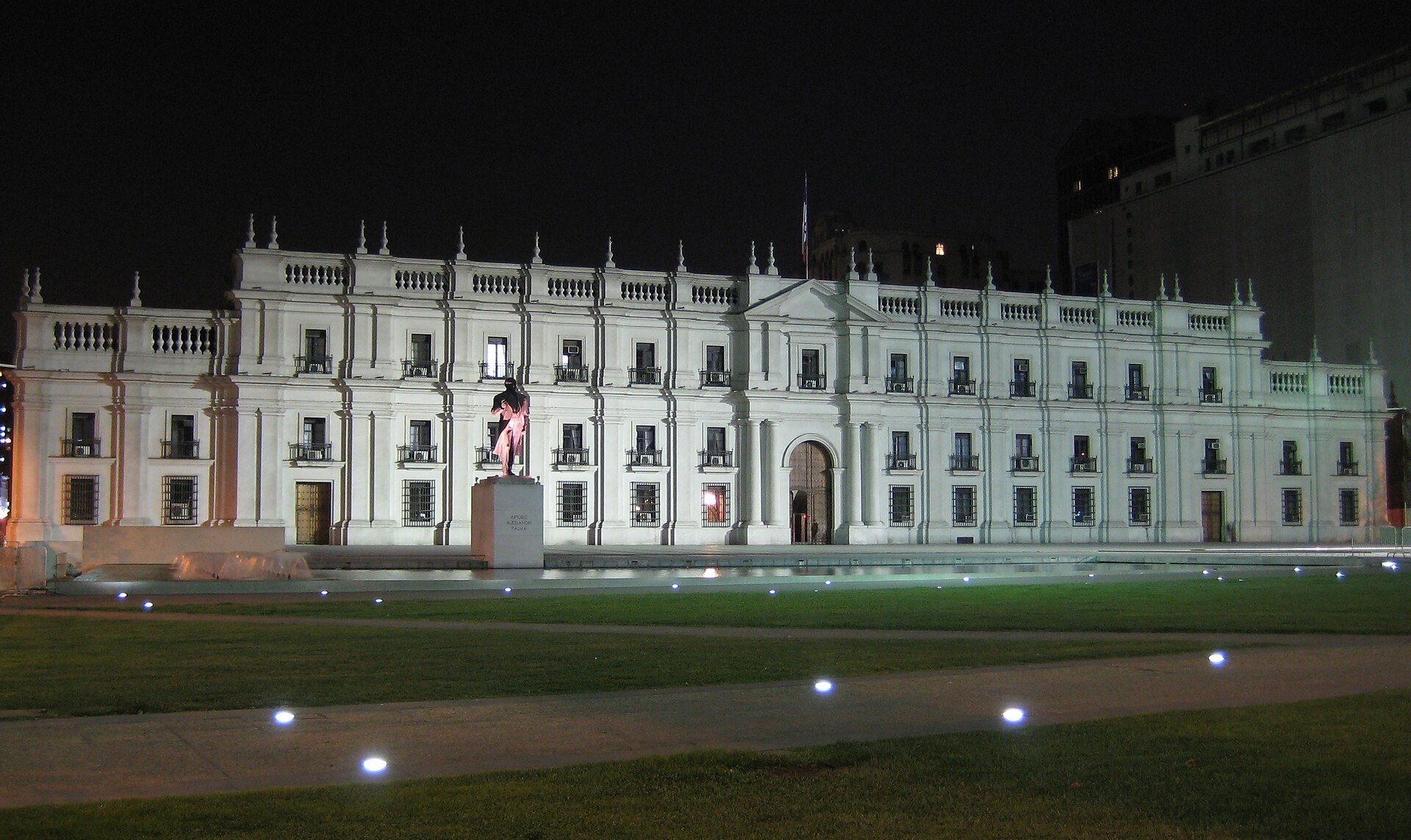 The Palacio de La Moneda in Santiago lit at night, with a statue on a plinth and lawn in the foreground