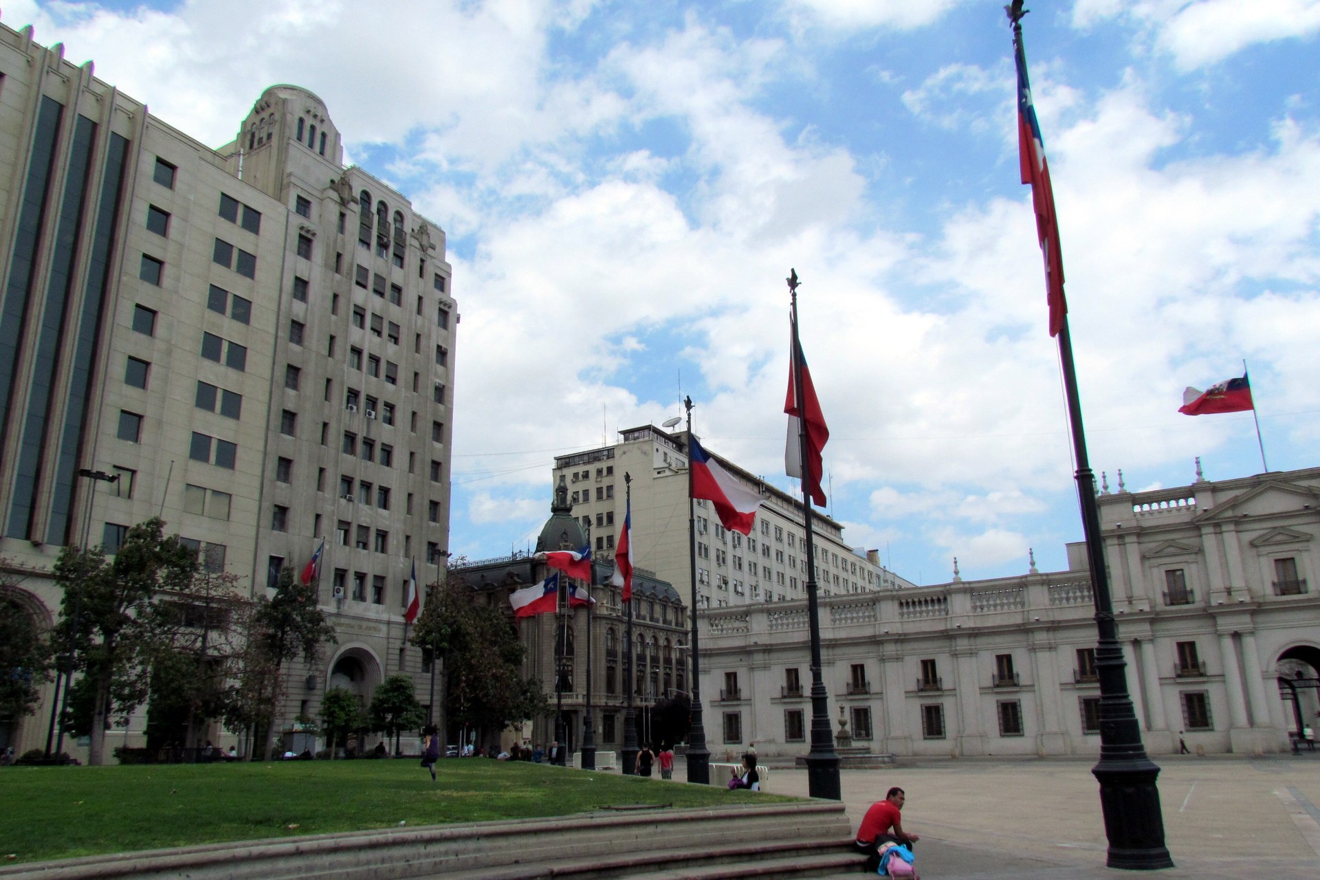Chilean flags on tall poles in the Plaza de la Constitución in Santiago, with the Palacio de La Moneda at right under a cloudy sky