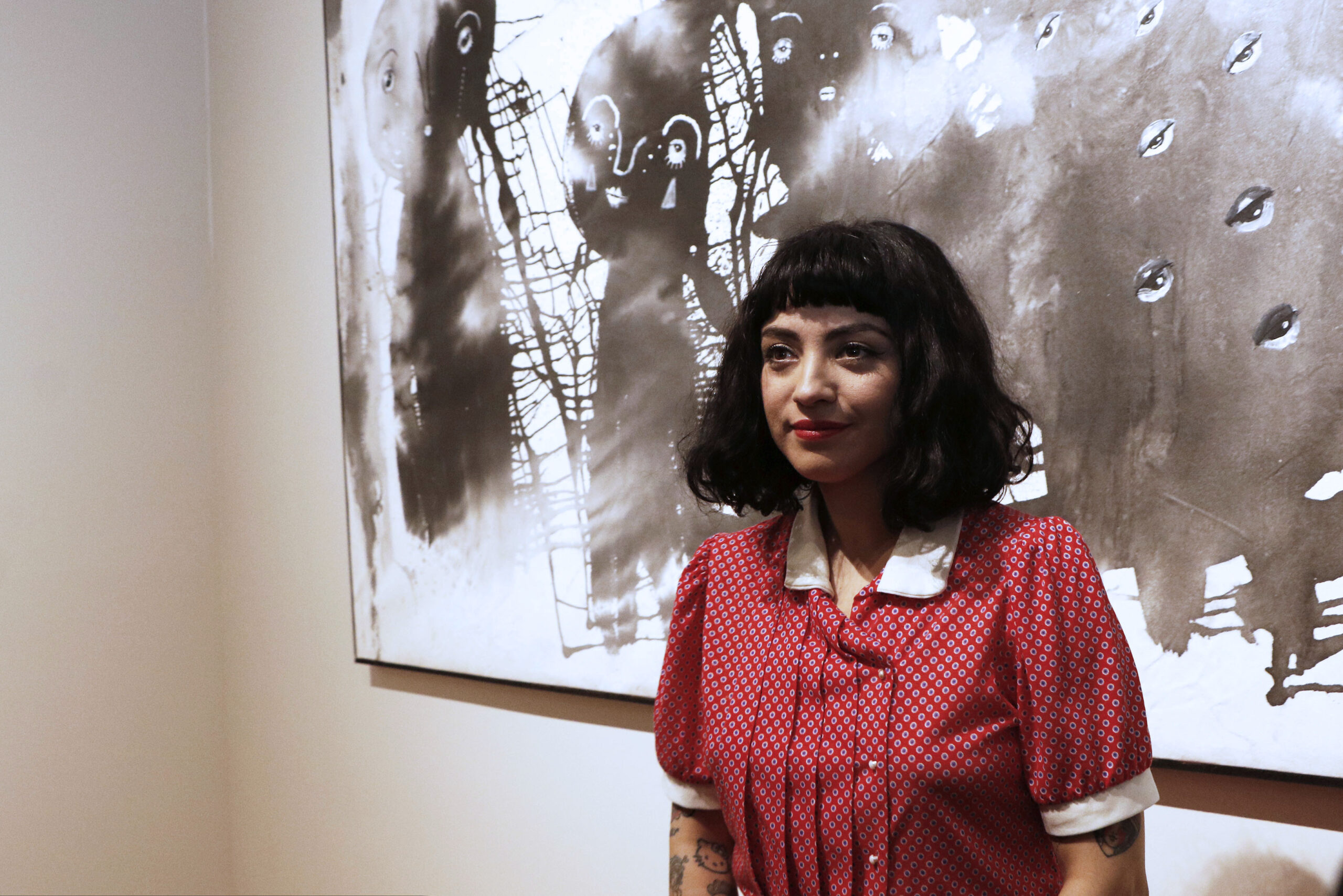 Singer Mon Laferte in a red patterned blouse standing in front of a large black-and-white painting
