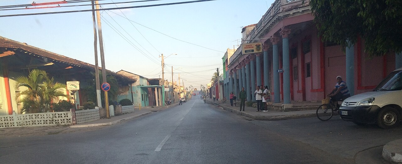 A wide street in Ciego de Ávila, Cuba, lined with colonnaded pink and blue buildings, with people waiting on the pavement and a man on a bicycle