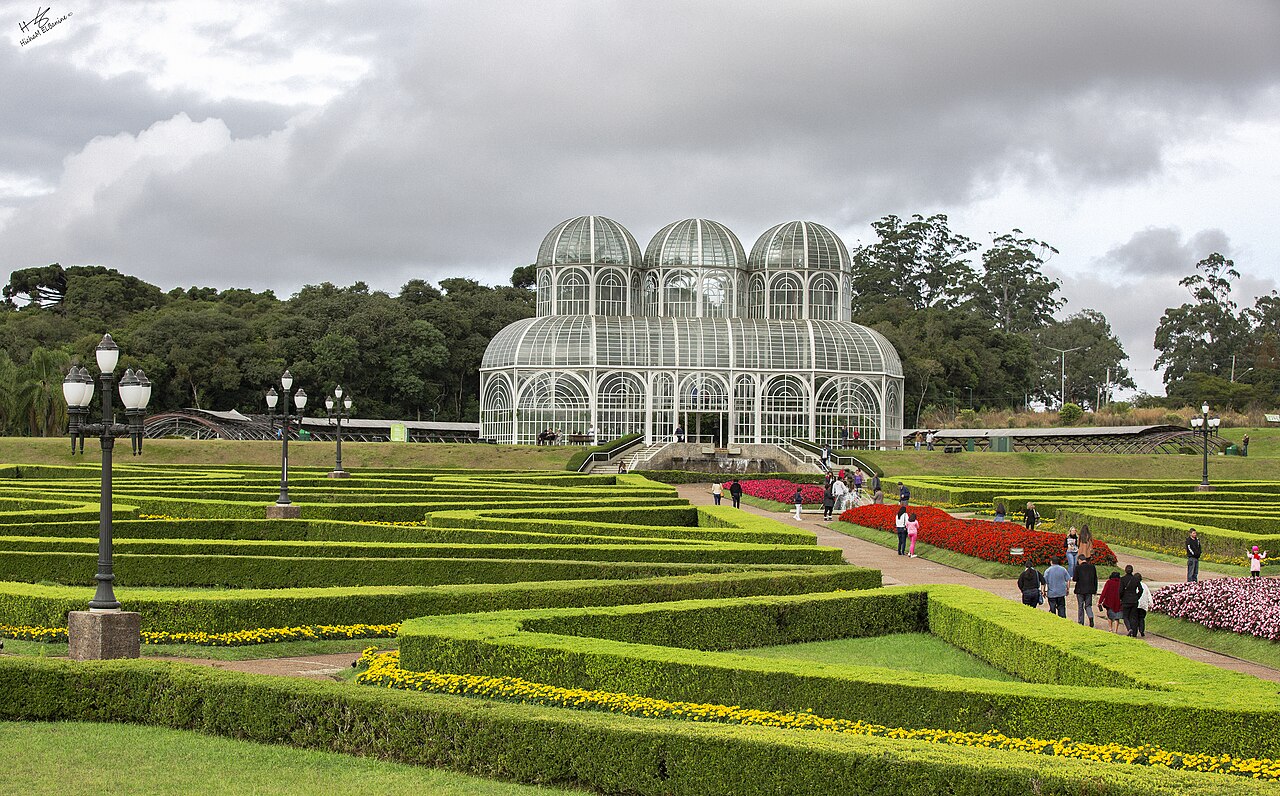 Glass greenhouse with three domes behind trimmed hedge gardens and red flower beds in Curitiba's Botanical Garden, with visitors on the paths.