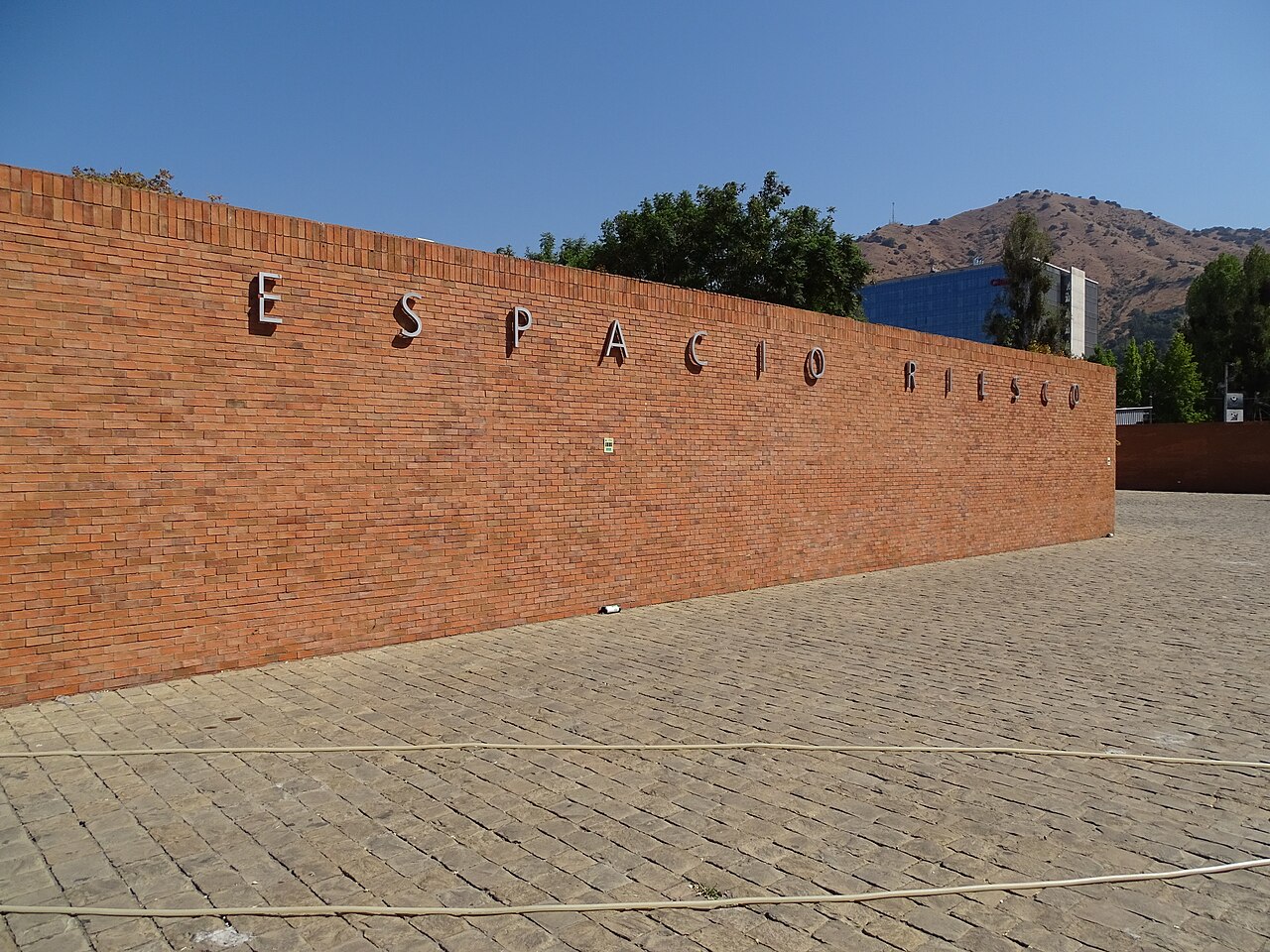 Long red-brick wall with the name Espacio Riesco in metal letters, above a cobbled forecourt, with a hill and trees behind under a clear blue sky.
