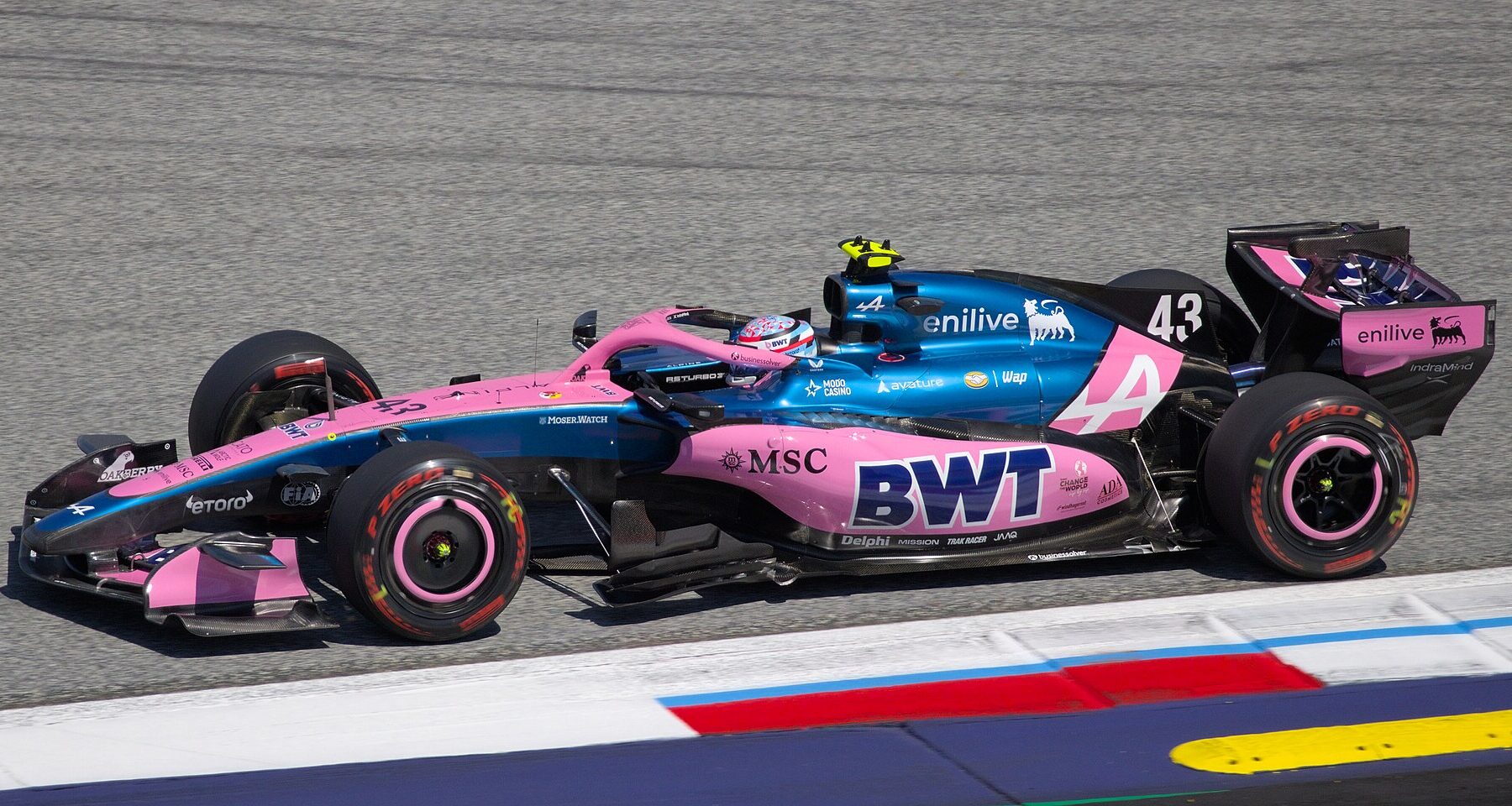 Side view of a blue and pink Alpine Formula 1 car with the number 43 driving past red, white and blue curbs on a grey asphalt track