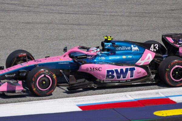 Side view of a blue and pink Alpine Formula 1 car with the number 43 driving past red, white and blue curbs on a grey asphalt track