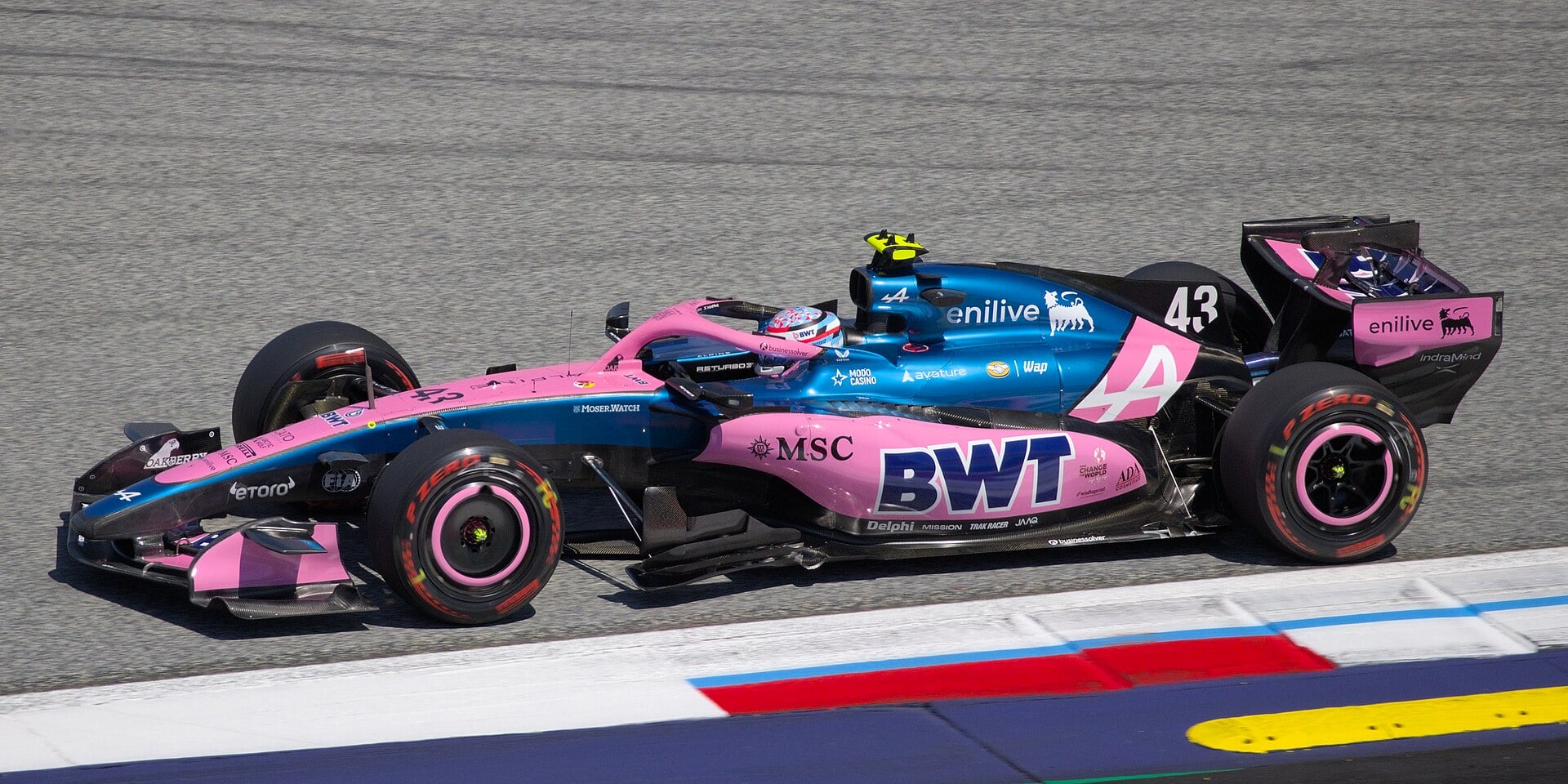 Side view of a blue and pink Alpine Formula 1 car with the number 43 driving past red, white and blue curbs on a grey asphalt track