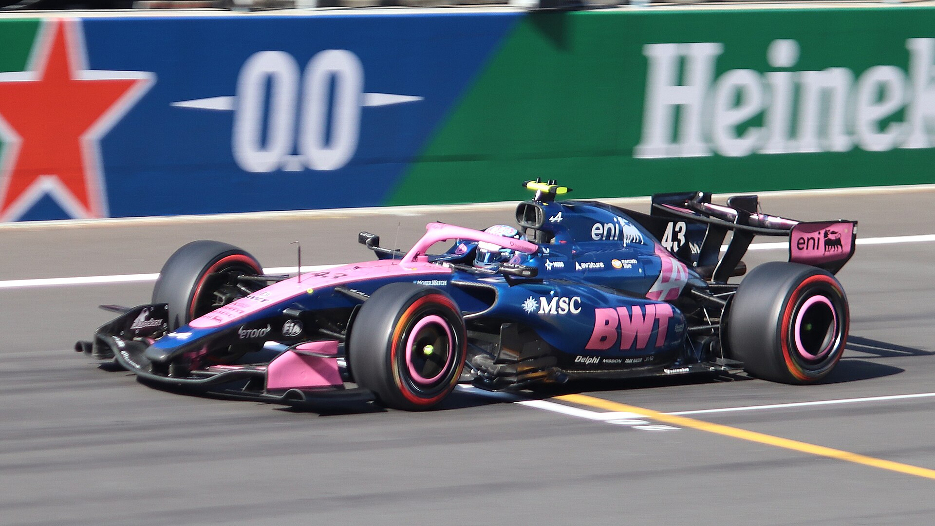 Front three-quarter view of a blue and pink Alpine Formula 1 car with the number 43 speeding past a green and blue advertising wall