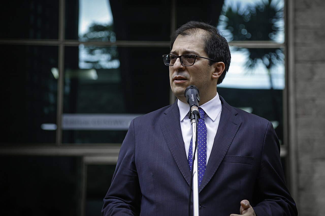 Francisco Barbosa, in glasses and a dark suit with a blue polka-dot tie, speaks into a microphone outside a glass-fronted building