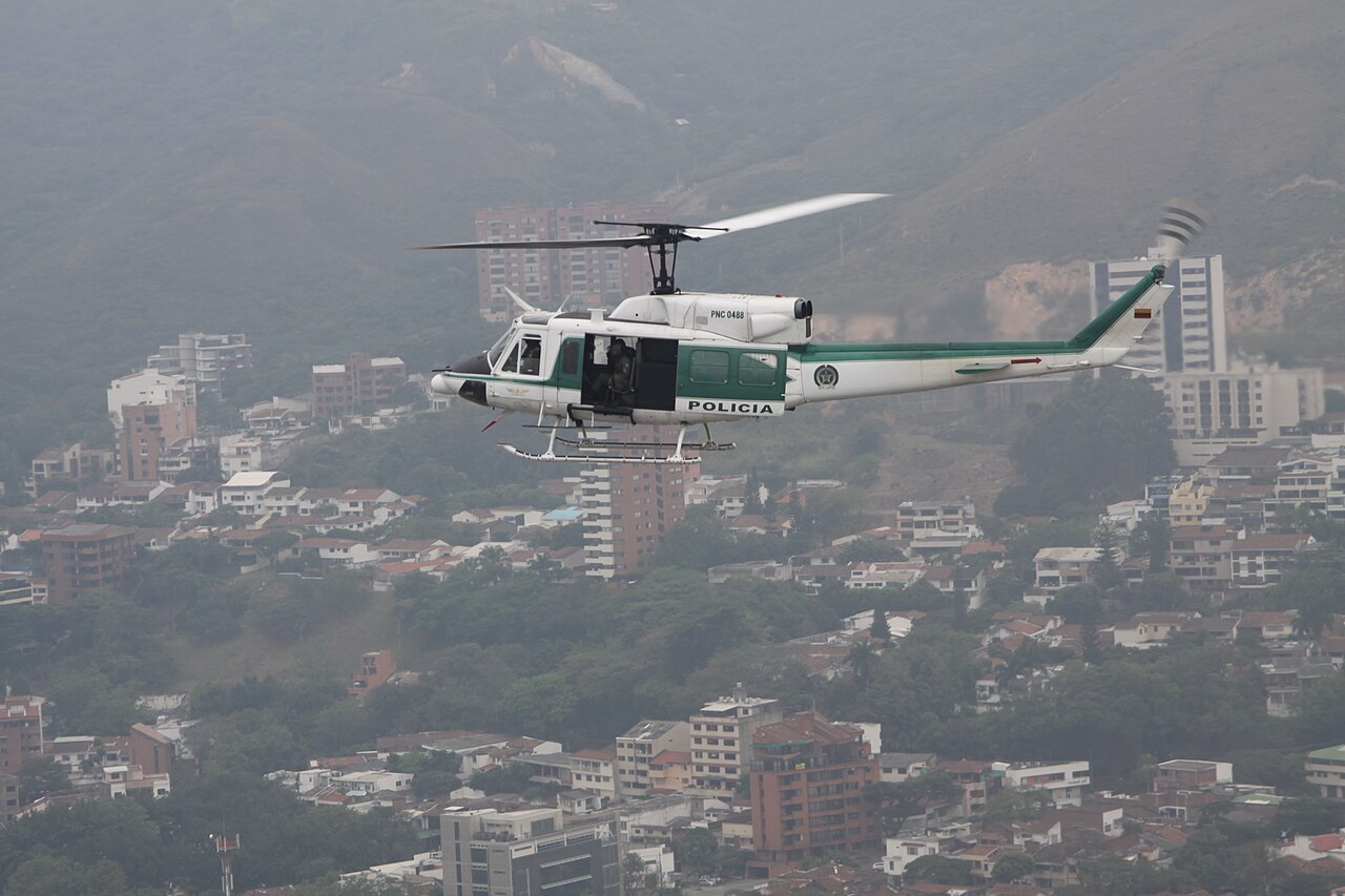 A white and green Colombian National Police helicopter flies over apartment blocks and hills in Cali