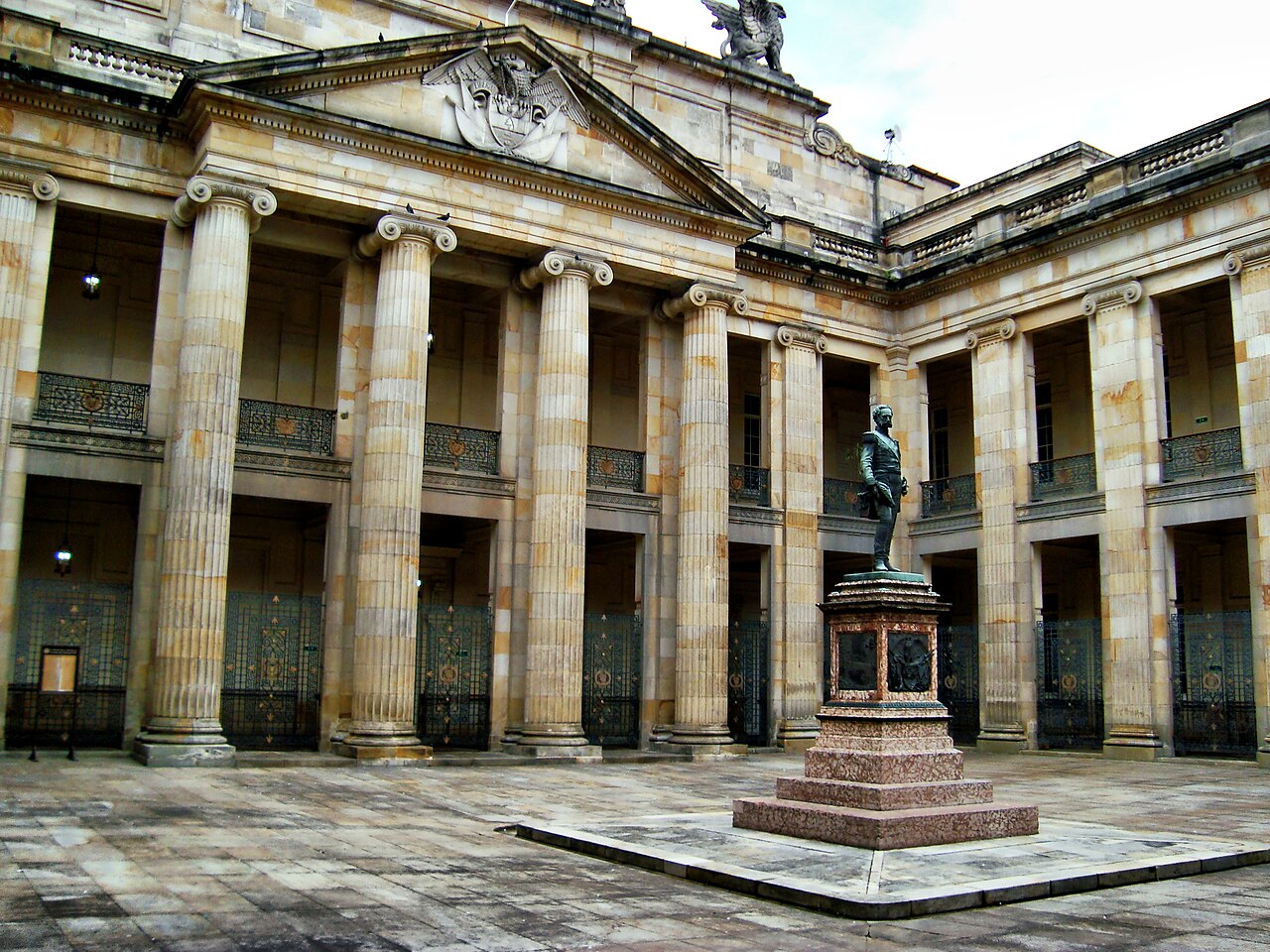 A stone courtyard with tall fluted columns, iron-grilled arches and a bronze statue on a pink granite pedestal