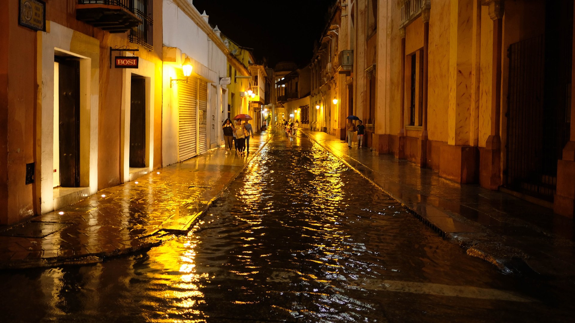 Night view of a flooded street between colonial buildings in Cartagena, Colombia, with people walking under umbrellas