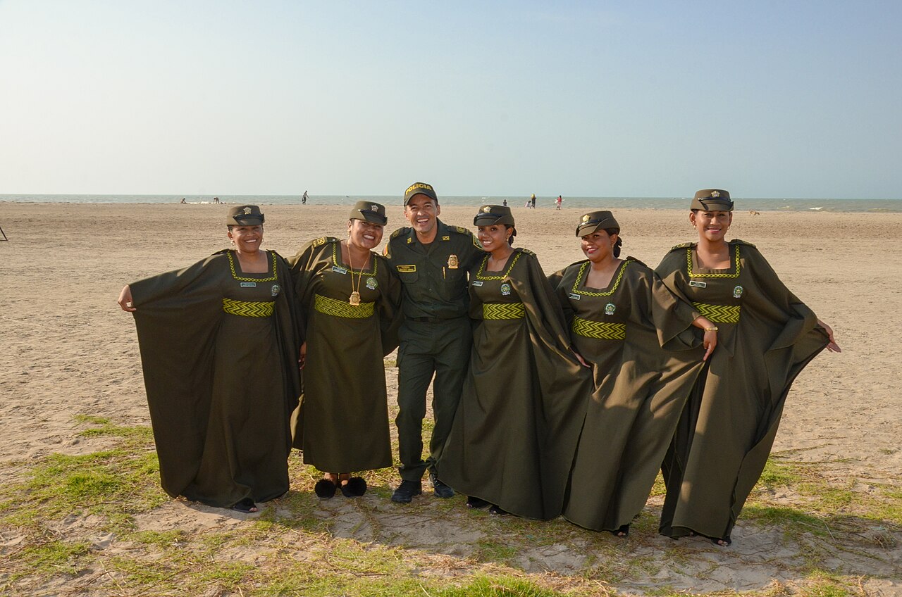 Five female officers in olive Wayuu-style robes and one male officer in uniform pose together on a sandy beach in La Guajira