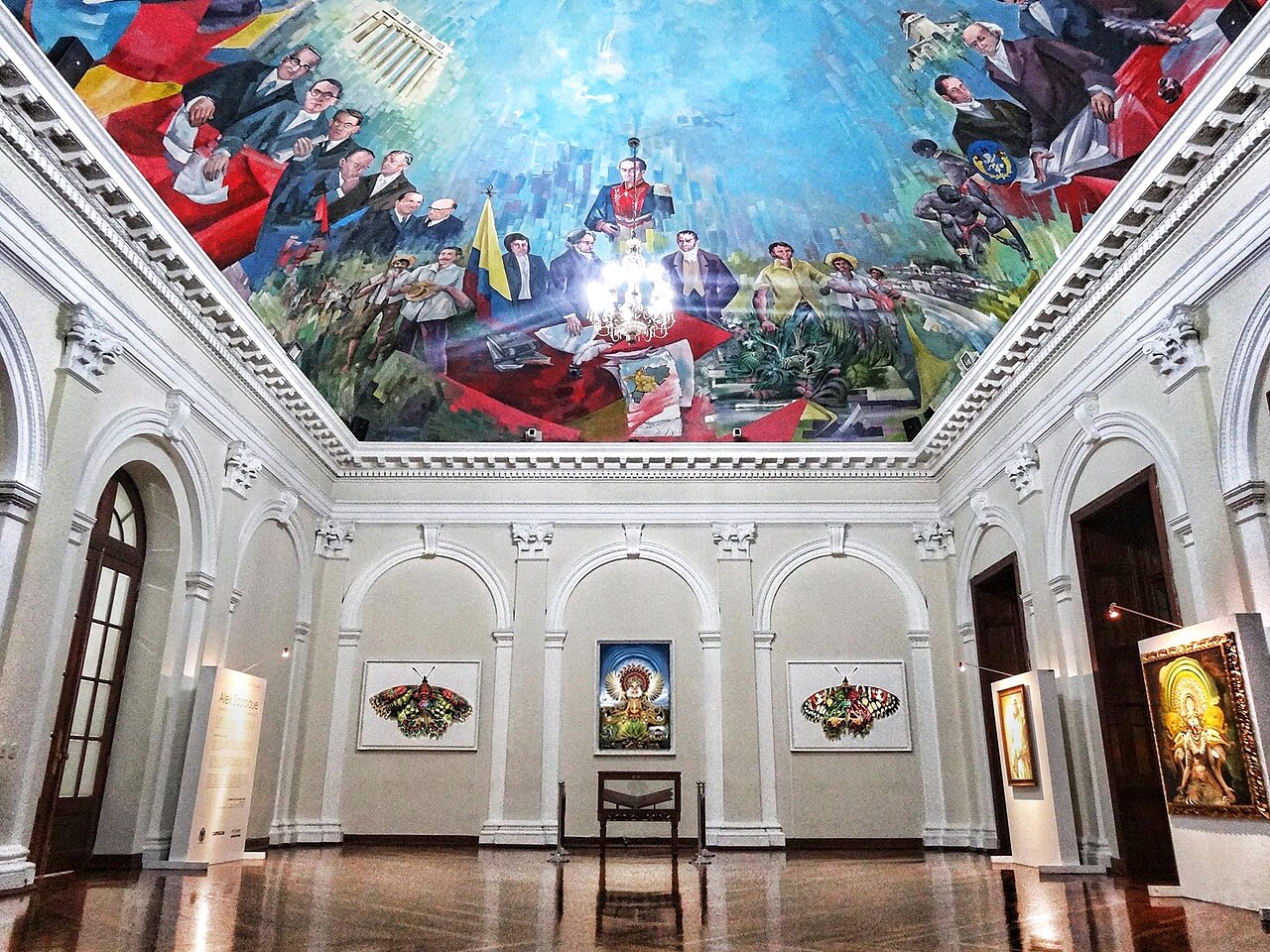 A grand hall with white arched walls, a ceiling painted with a colourful mural of historical figures and a chandelier, and framed artworks on the walls