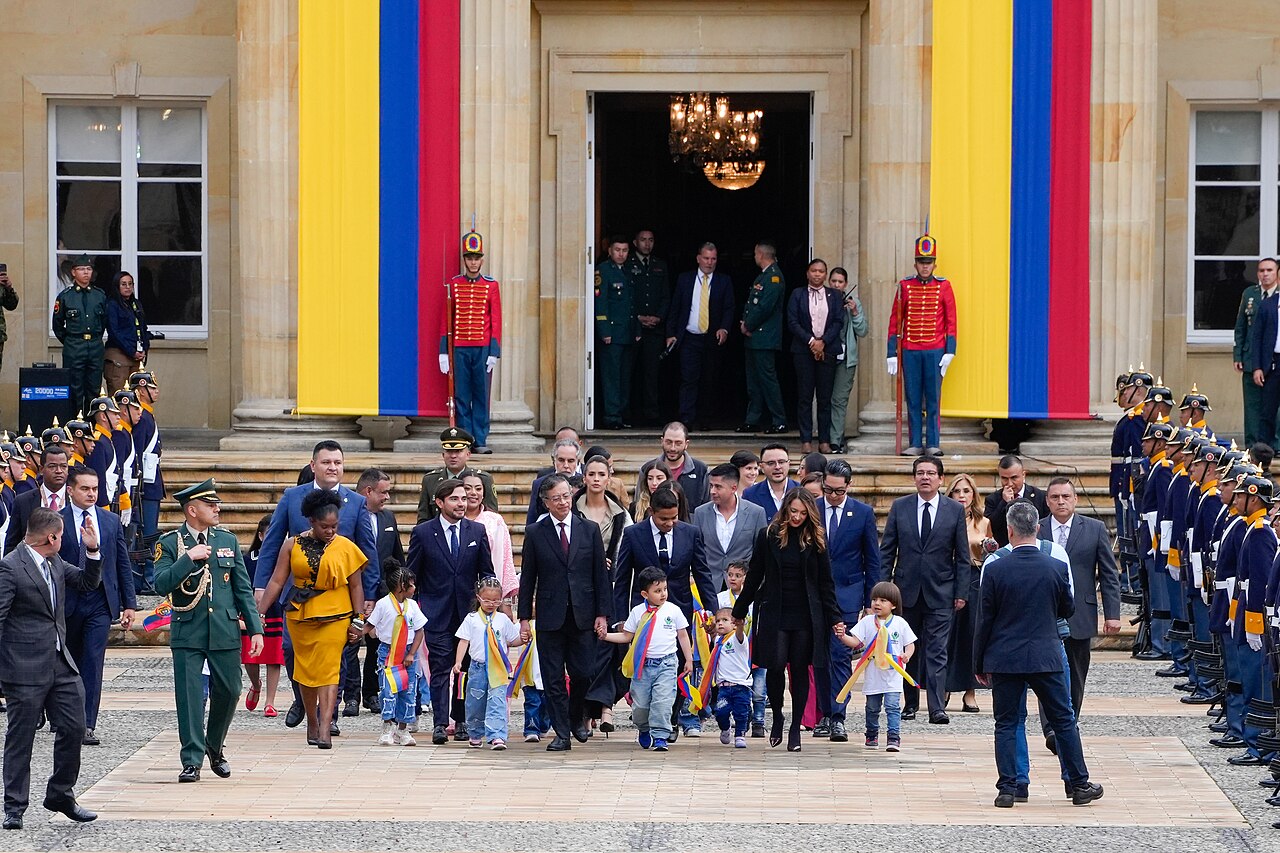 Officials, children and uniformed honour guards in front of a stone building hung with two large yellow, blue and red Colombian flags