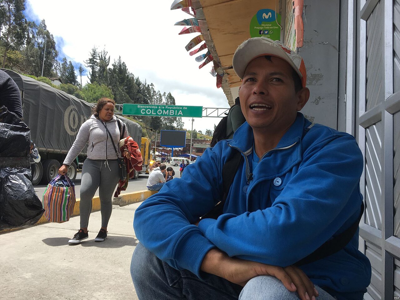 A man in a blue jacket and cap sits outside a shop as a woman walks past with bags, a Welcome to the Republic of Colombia sign behind them