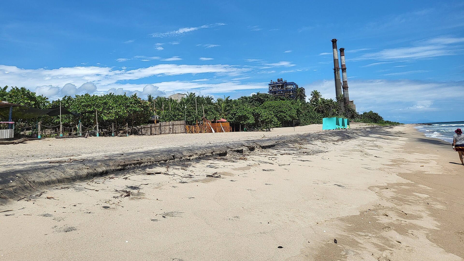 Two tall striped chimneys and a blue boiler building of a thermal power station behind palm trees on a beach in La Guajira, Colombia