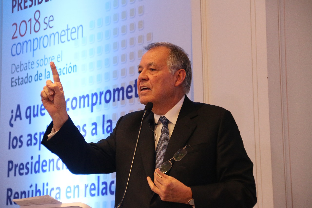 A grey-haired man in a dark suit speaks at a microphone with one finger raised, in front of a blue screen with Spanish text