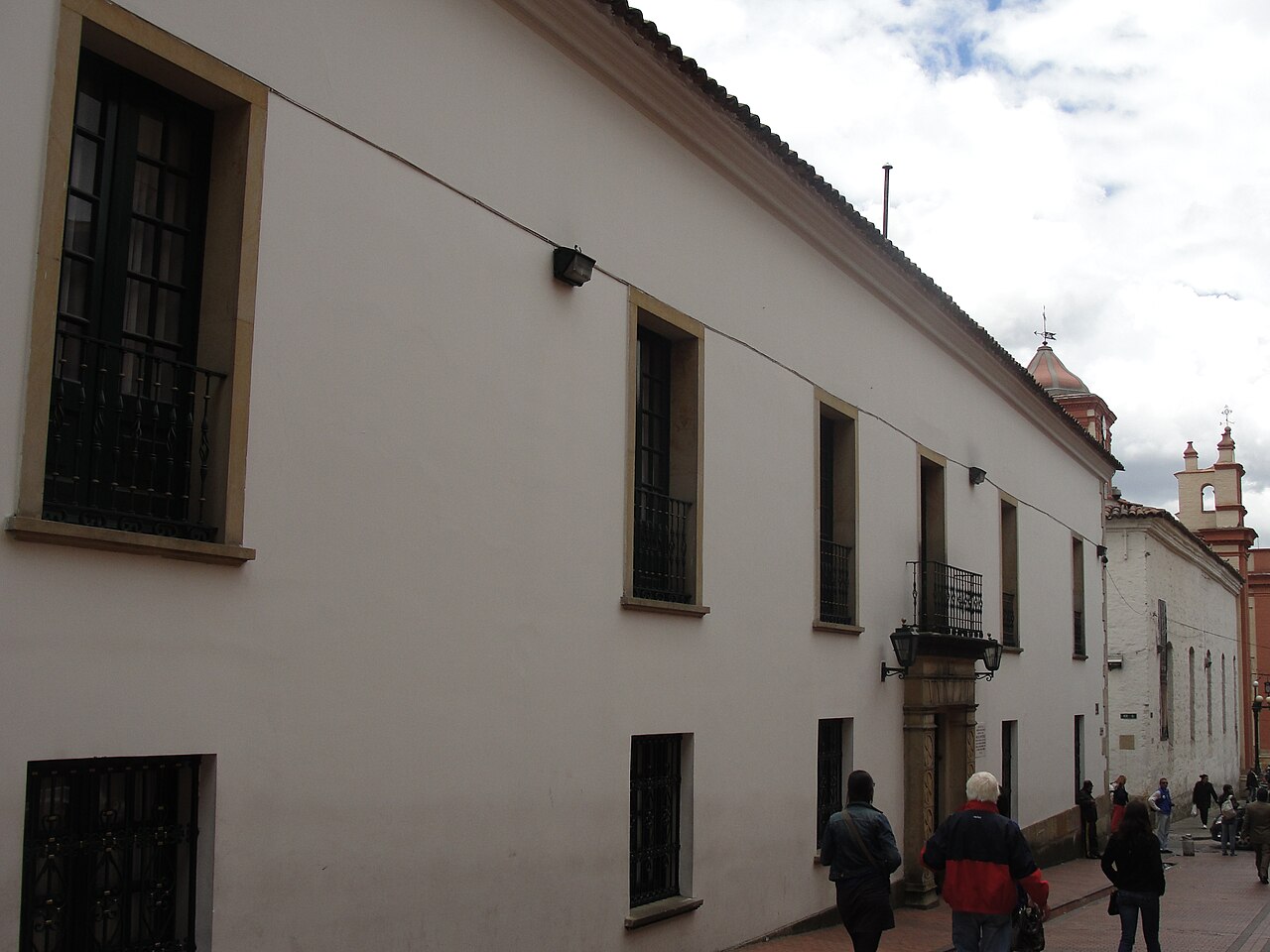 Long white colonial facade of the Palacio de San Carlos in Bogotá with barred windows, iron balconies and pedestrians on the street