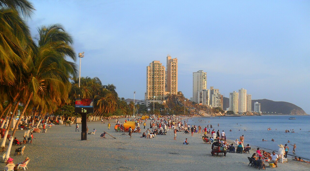 Palm-lined El Rodadero beach in Santa Marta with sunbathers, high-rise hotels and a green headland