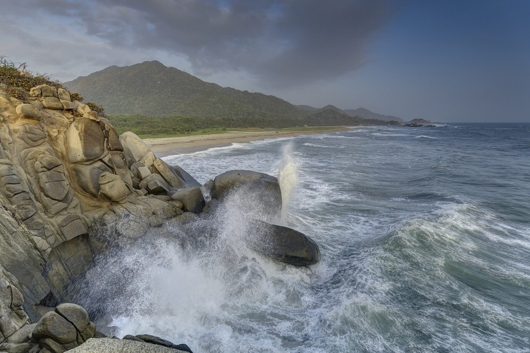 Waves breaking on granite boulders beside a beach and forested hills in Tayrona National Park near Santa Marta