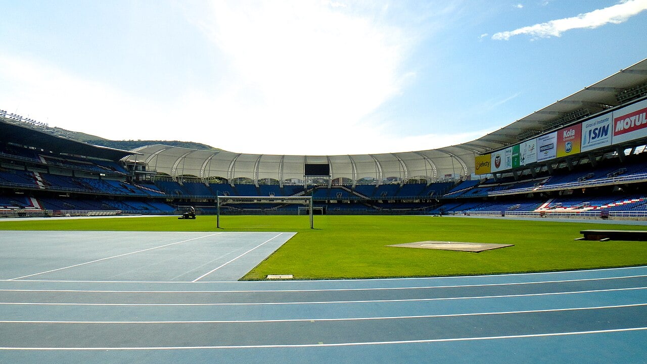 Inside the Estadio Olímpico Pascual Guerrero in Cali, with blue stands, a white roof, green pitch and a blue running track under a bright sky.