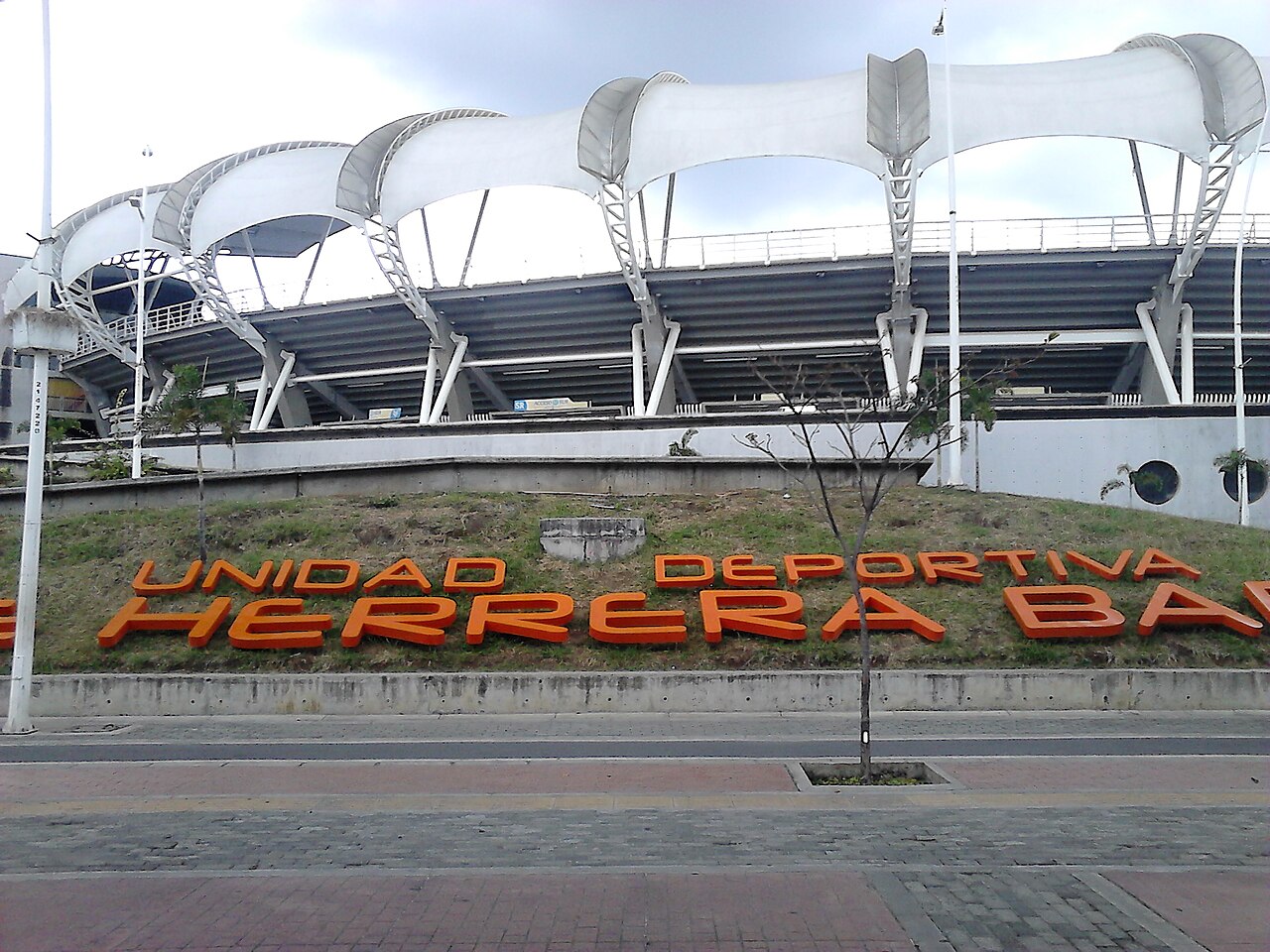 The white fabric roof and concrete stands of the Estadio Olímpico Pascual Guerrero in Cali, with orange letters on a grass bank in front.