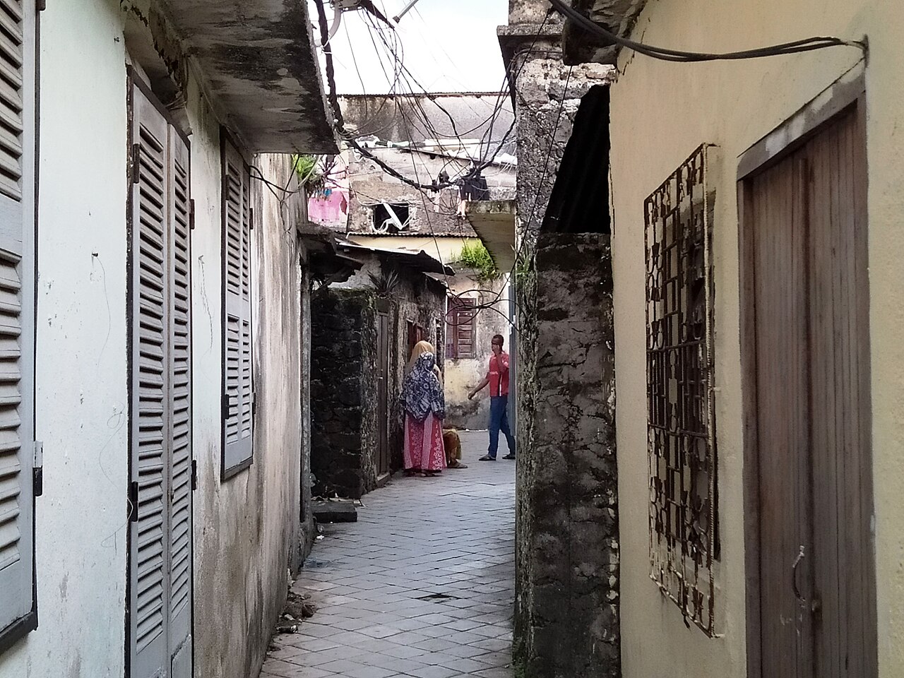 A narrow paved alley between old stone houses in the medina of Moroni, the capital of Comoros, with two people talking at the far end