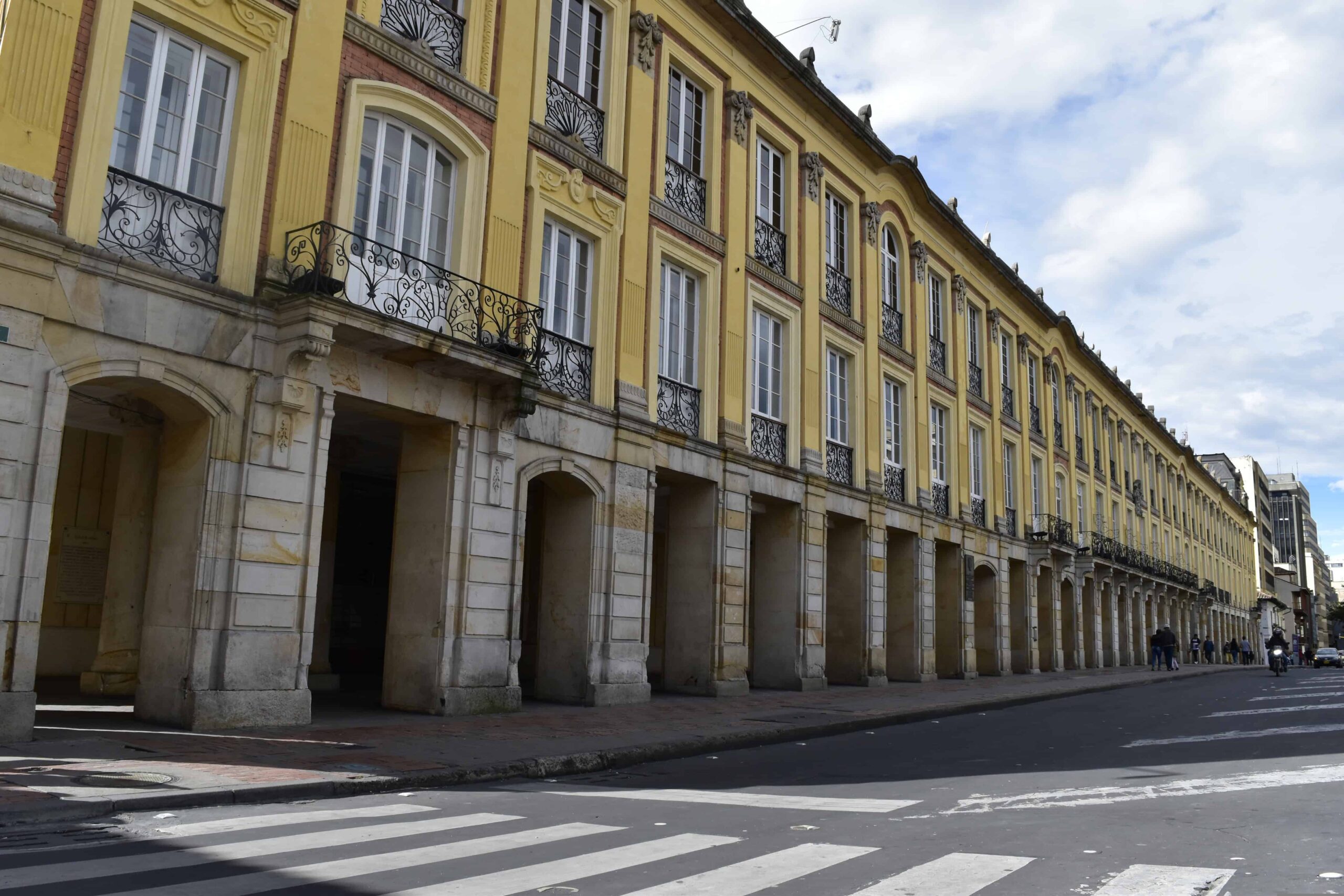 A long yellow colonial-style building with arched ground-floor arcades and iron balconies beside a street in central Bogotá.